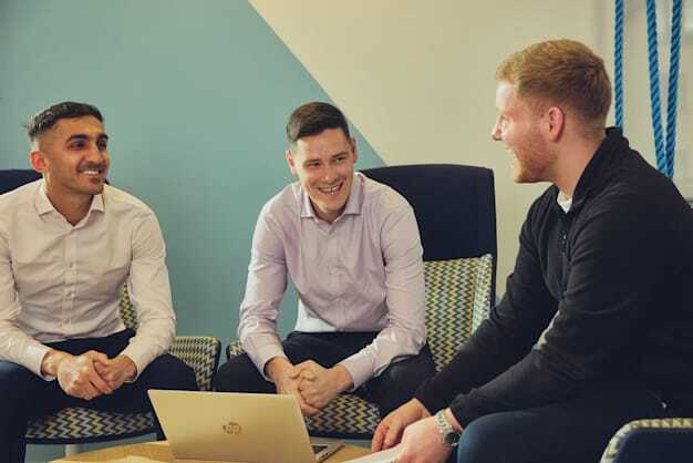 Three SmartSearch colleagues seated in office chairs around a laptop, smiling during a casual meeting