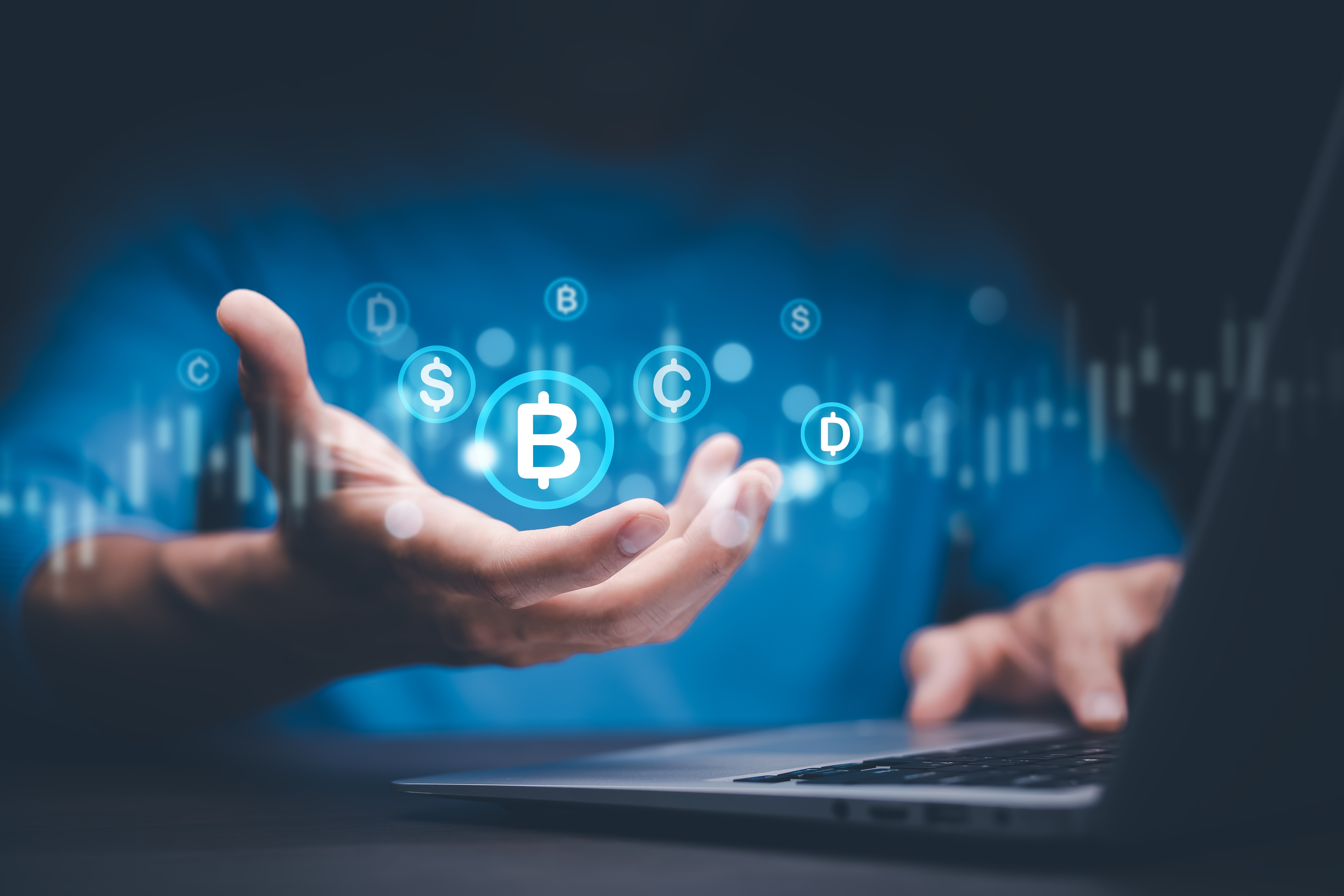 Male hand holding up various cryptocurrencies in front of a laptop